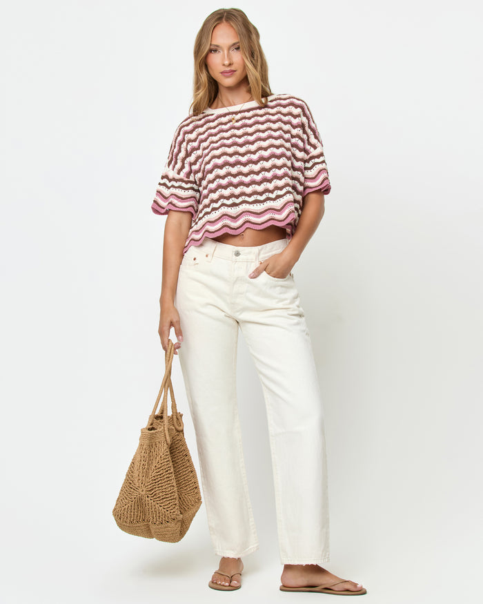 Front view on model of the LSPACE Sunchaser Top in Neapolitan, a short-sleeve crochet top in a brown, pink, and white wave pattern with scalloped hem, styled with white wide-leg jeans and a straw tote bag.