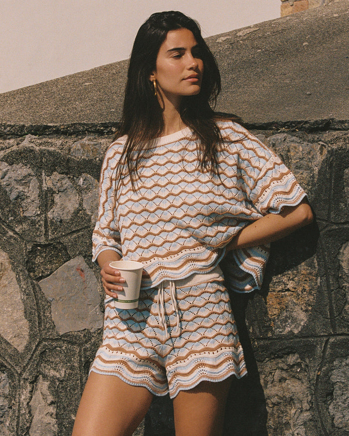 Angled view of a model wearing the LSPACE Sunchaser Top in north shores, a boxy crochet knit top with short sleeves and a wave scallop pattern in light blue and tan, photographed outdoors, styled with the matching shorts.