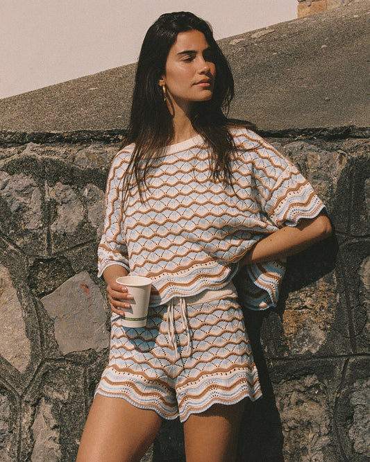 Angled view of a model wearing the LSPACE Sunchaser Top in north shores, a boxy crochet knit top with short sleeves and a wave scallop pattern in light blue and tan, photographed outdoors, styled with the matching shorts.