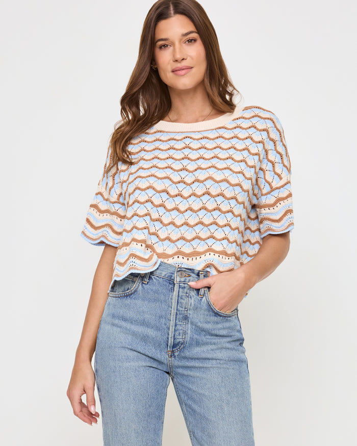 Front view of a model wearing the LSPACE Sunchaser Top in north shores, a relaxed-fit crochet knit top with a round neckline, short sleeves, and a wave scallop pattern in light blue and tan, styled with blue denim jeans.