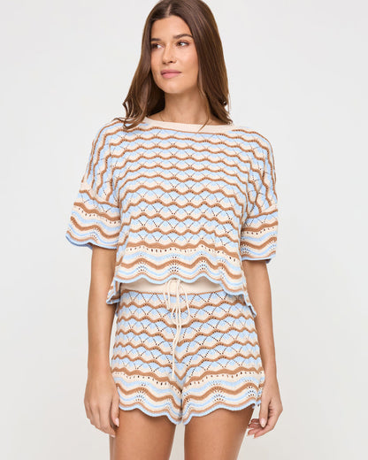 Angled front view of a model wearing the LSPACE Sunchaser Top in north shores, a boxy crochet knit top with a wave scallop pattern in light blue and tan, paired with the coordinating shorts.