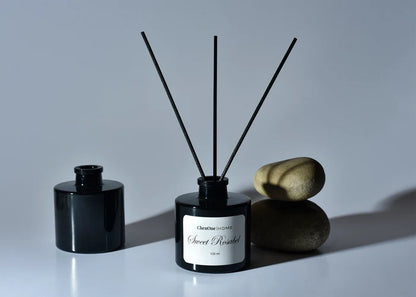 Sweet Rosabel reed diffuser with black bottle and four dark reeds, styled with stones.