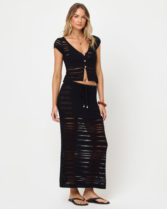 Side view of model wearing the LSPACE Tula Top in black, a semi-sheer crochet cropped top with cap sleeves with the mathcing Skirt.