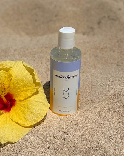 The Undershower Swimwear Detergent bottle displayed on sand beside a yellow hibiscus flower.