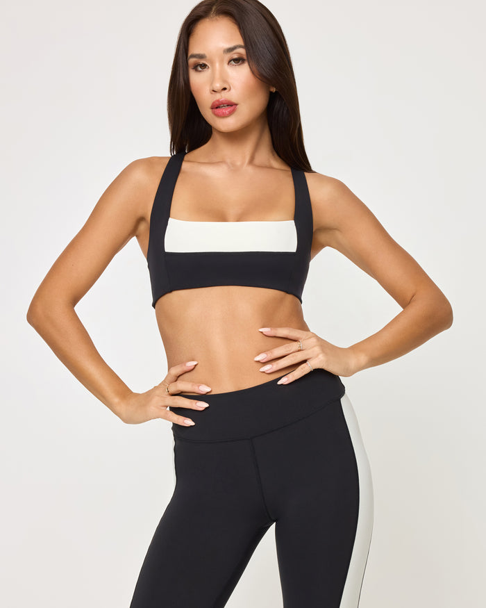 Lifestyle editorial shot of a model wearing the LSPACE Uppercut Bra in black-cream paired with matching black-cream high-rise active pants, in an outdoor setting.