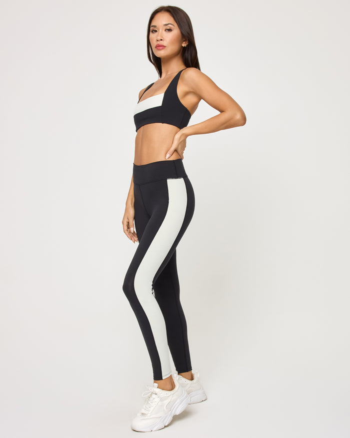 Side view of the LSPACE Uppercut Bra in black-cream on a model against a white background, paired with black-cream high-rise active pants, showing the profile of the matching outfit.